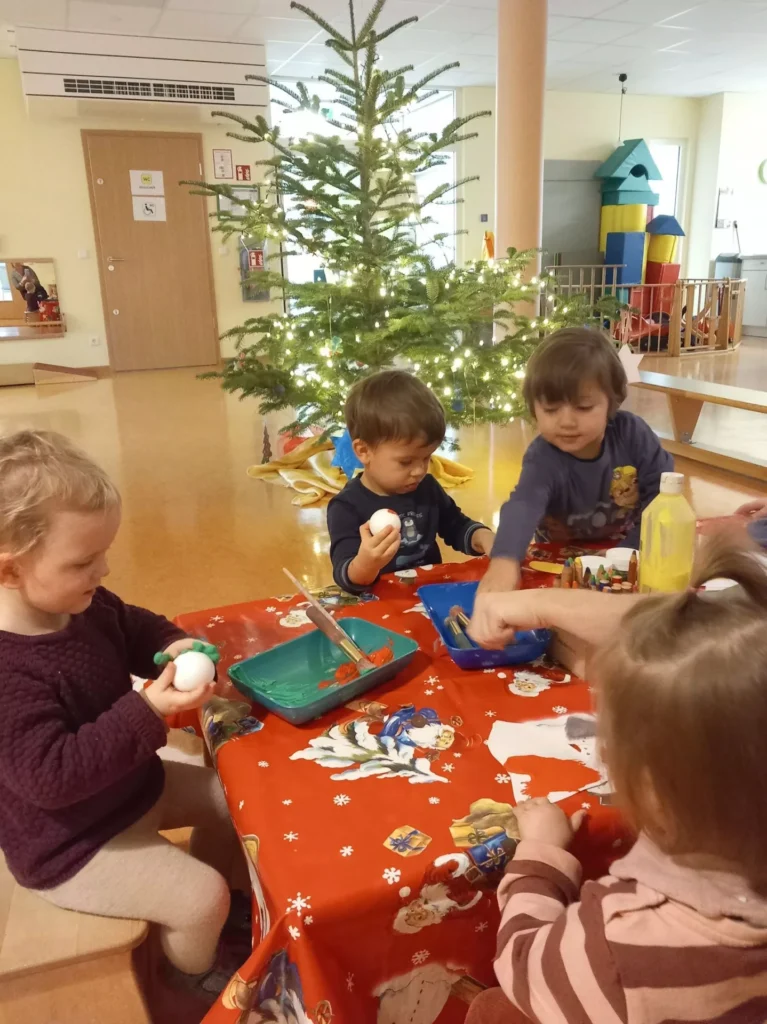 Auf diesem Bild sind vier Kinder an einem Tisch zu sehen, die etwas basteln. Im Hintergrund ist der Weihnachtsbaum zu erkennen.